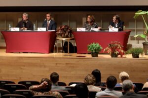 Four people sit behind two tables on stage, two person per table, as part of a panel.
